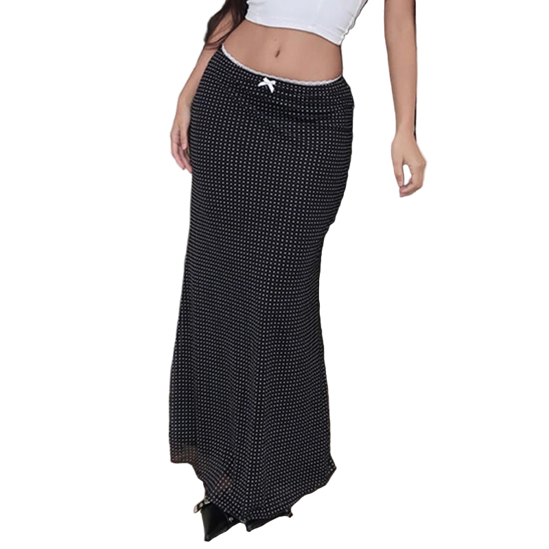 A woman wearing a white top and a black ankle-length mesh skirt with polka dots and a bow detail, standing by a door.